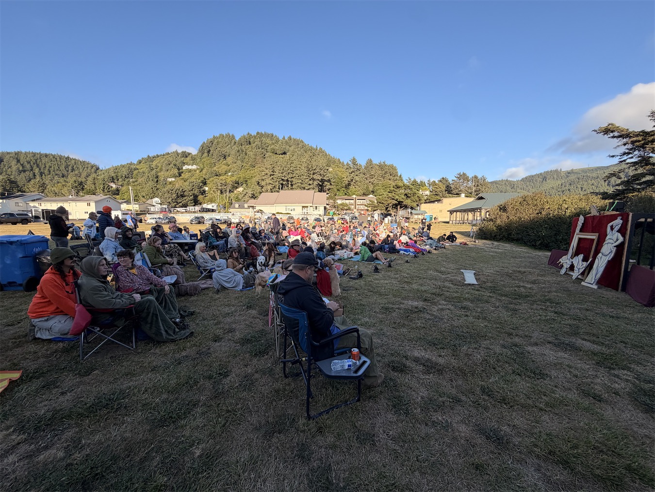 An audience on blankets and camp chairs watches on a grassy field in Yachats at golden hour, with wooded coastal hills behind and the painted opera-house stage at right.