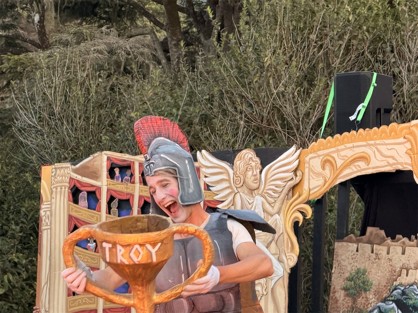 The fly actor, in a crested Roman helmet, laughs while hoisting a large golden cup labeled TROY beside a carved angel and a miniature puppet stage.