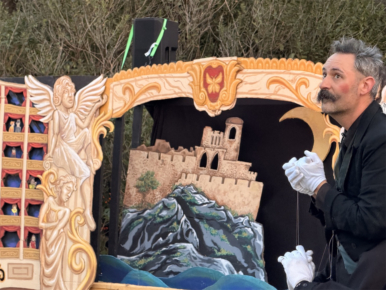 Close-up of the fly actor in profile, white-gloved hands raised as if cleaning them, beside the golden proscenium and a painted castle backdrop.