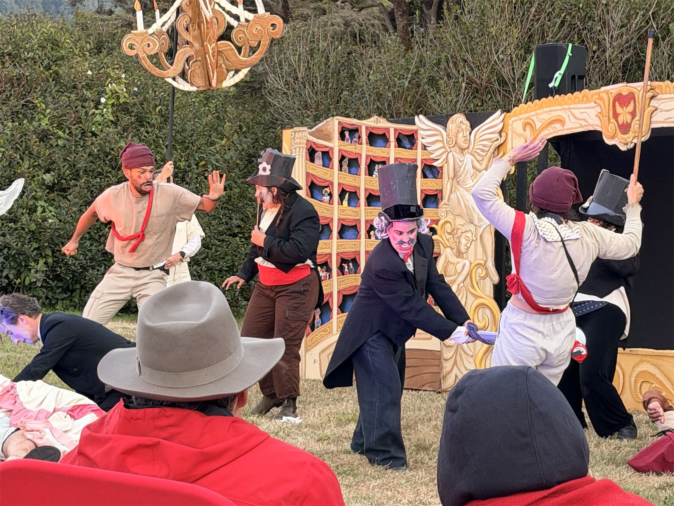 Costumed performers in top hats stage a duel in front of the miniature opera stage, one lunging with a blade beneath a hanging chandelier.