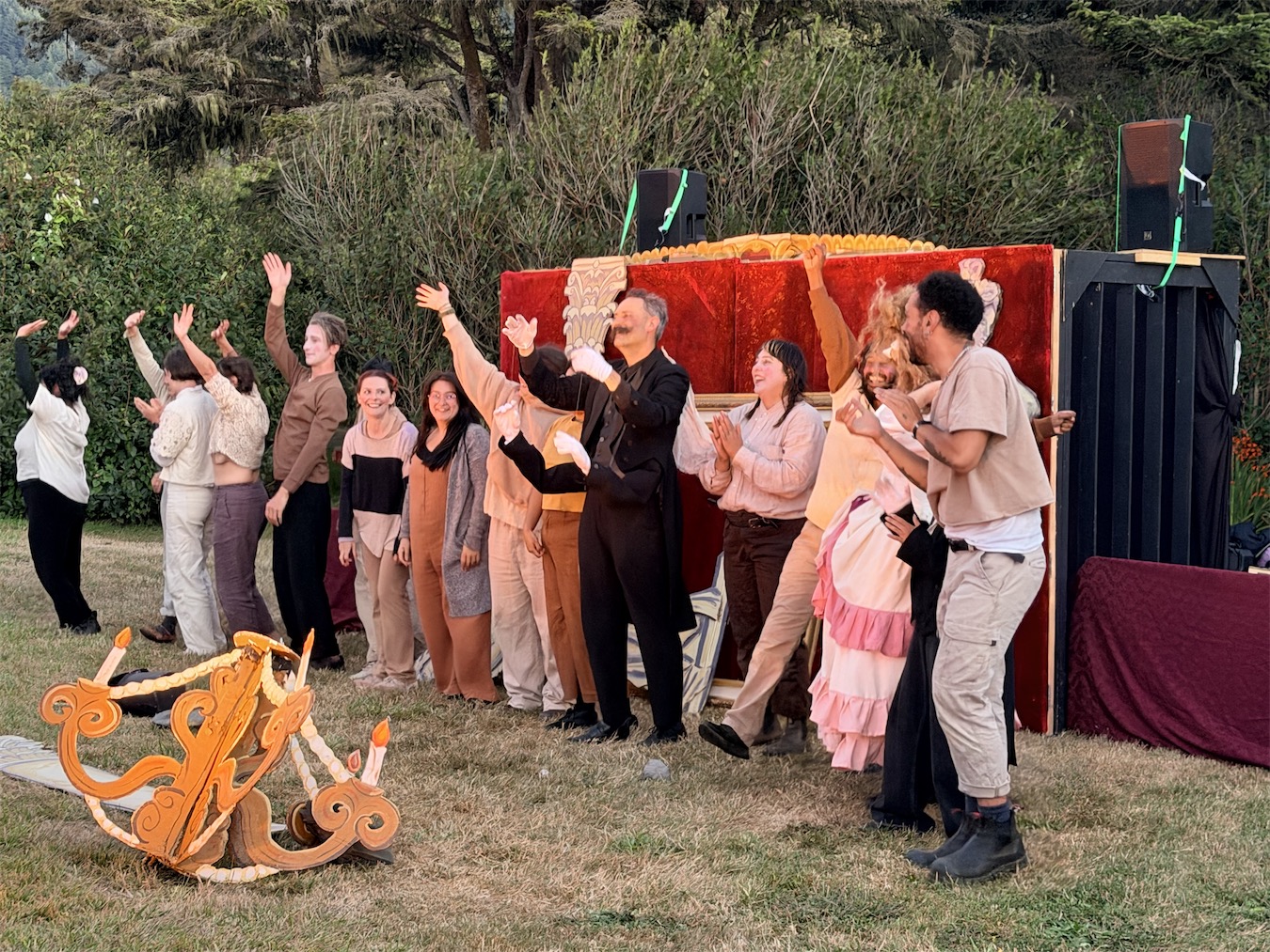 The String and Shadow company lines up with arms raised for a curtain call in front of the red-curtained stage as the sun sets over the hills.