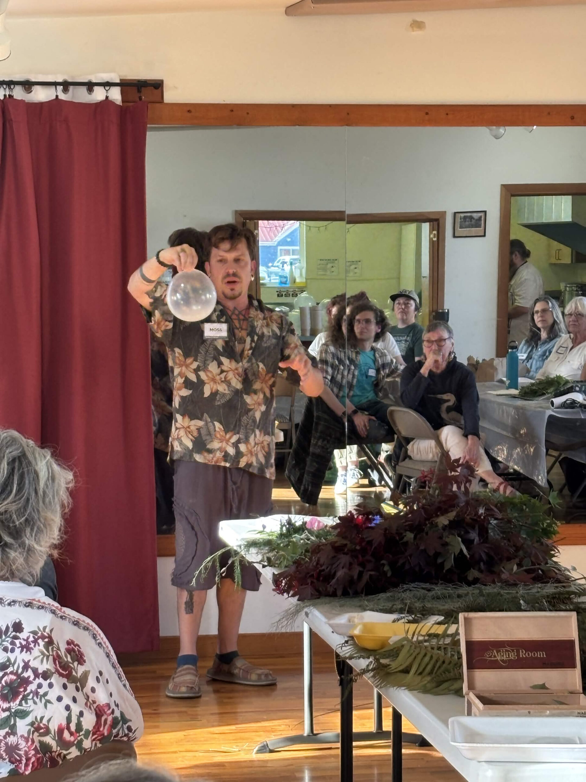 Moss Webster, in a floral shirt and name tag, holds up a balloon for the seated group while demonstrating at the front of the room, with tables of foraged ferns and dark foliage in front of him.