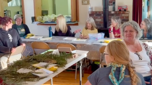 Participants seated around a table laughing together, sprigs of fern and greenery laid out on the paper-covered tables in front of them.