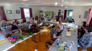 A wide view of the community hall with participants seated at long tables working with foraged greenery and flowers, LED string lights on the front table.