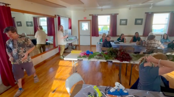 Participants at long tables spread with foraged ferns, branches, and flowers in the sunlit community hall during the lantern workshop.