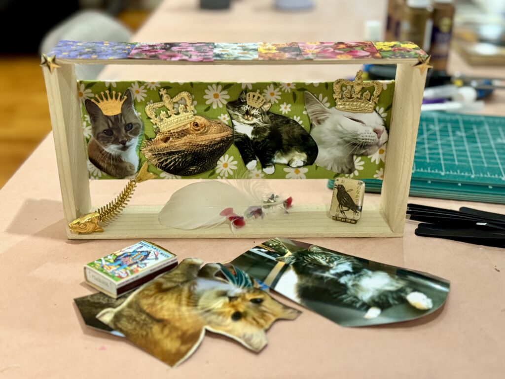 A wooden shadow box holding cutout photos of three cats and a bearded dragon, each wearing a small gold crown, against green daisy fabric, with a gold fish skeleton and a white feather below.