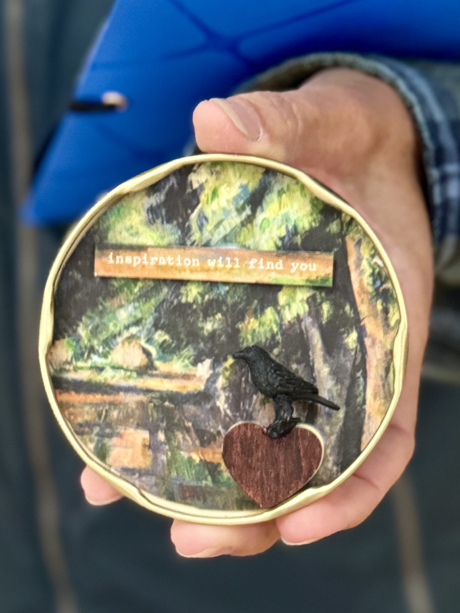 A hand holding a small round tin lid with a painted forest landscape, a black raven figure standing on a wooden heart, and a typed strip reading inspiration will find you.