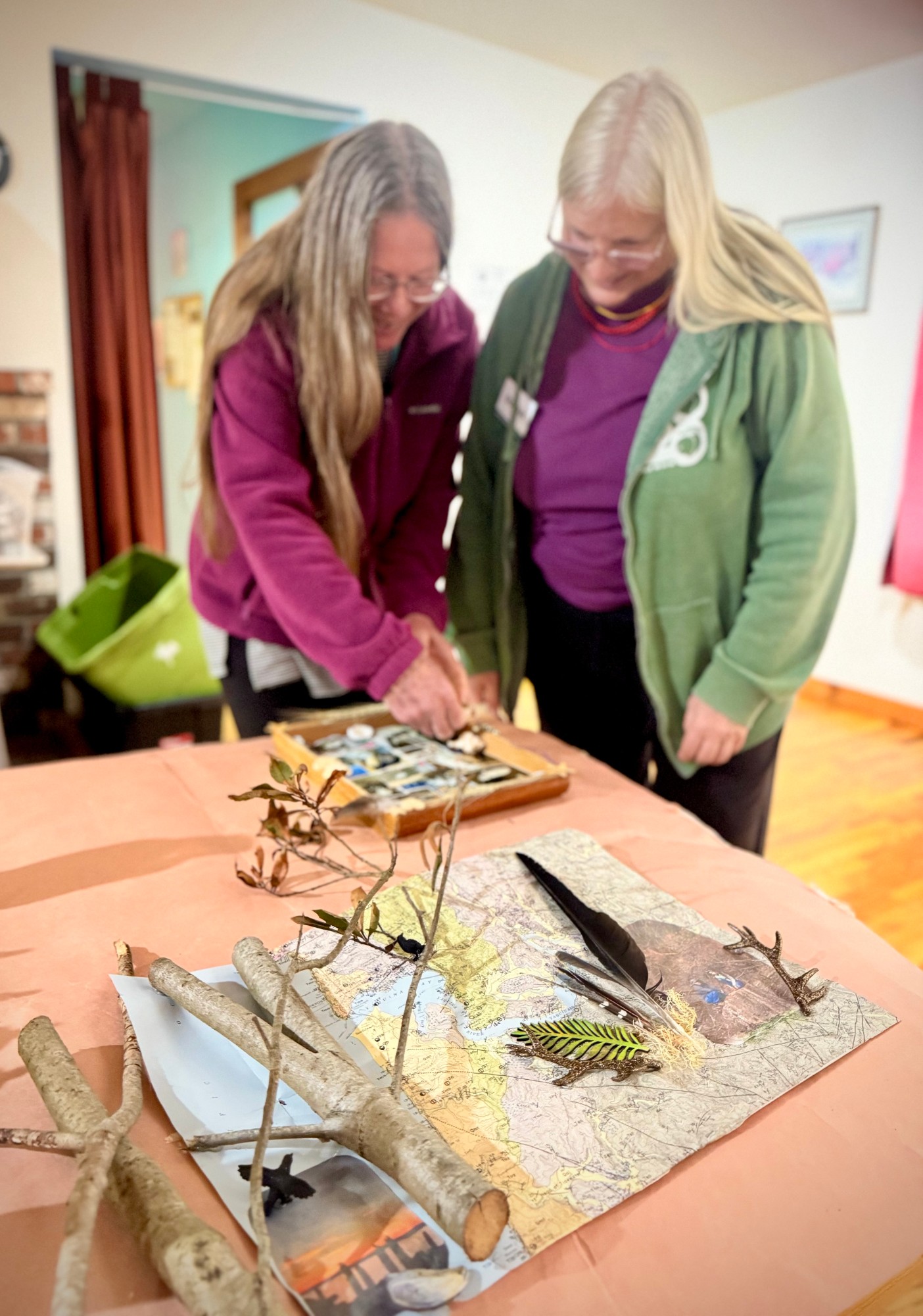 A coastal map laid under driftwood branches, a black feather, and a sprig of fern, with a participant arranging a box of small items behind it.