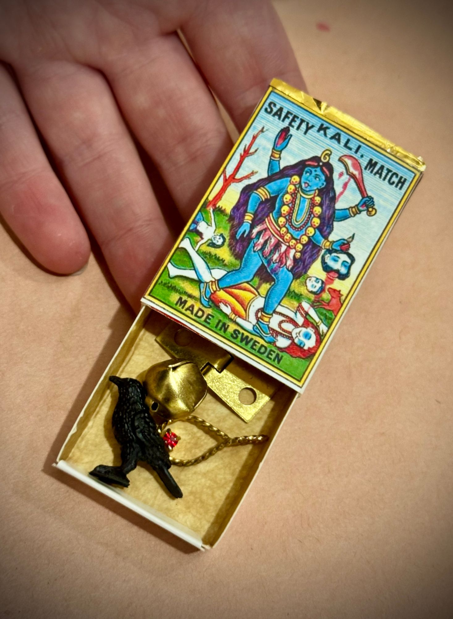 An open matchbox holding a small black raven figure, a brass bell, a brass hinge, and a gold chain with a red jewel.