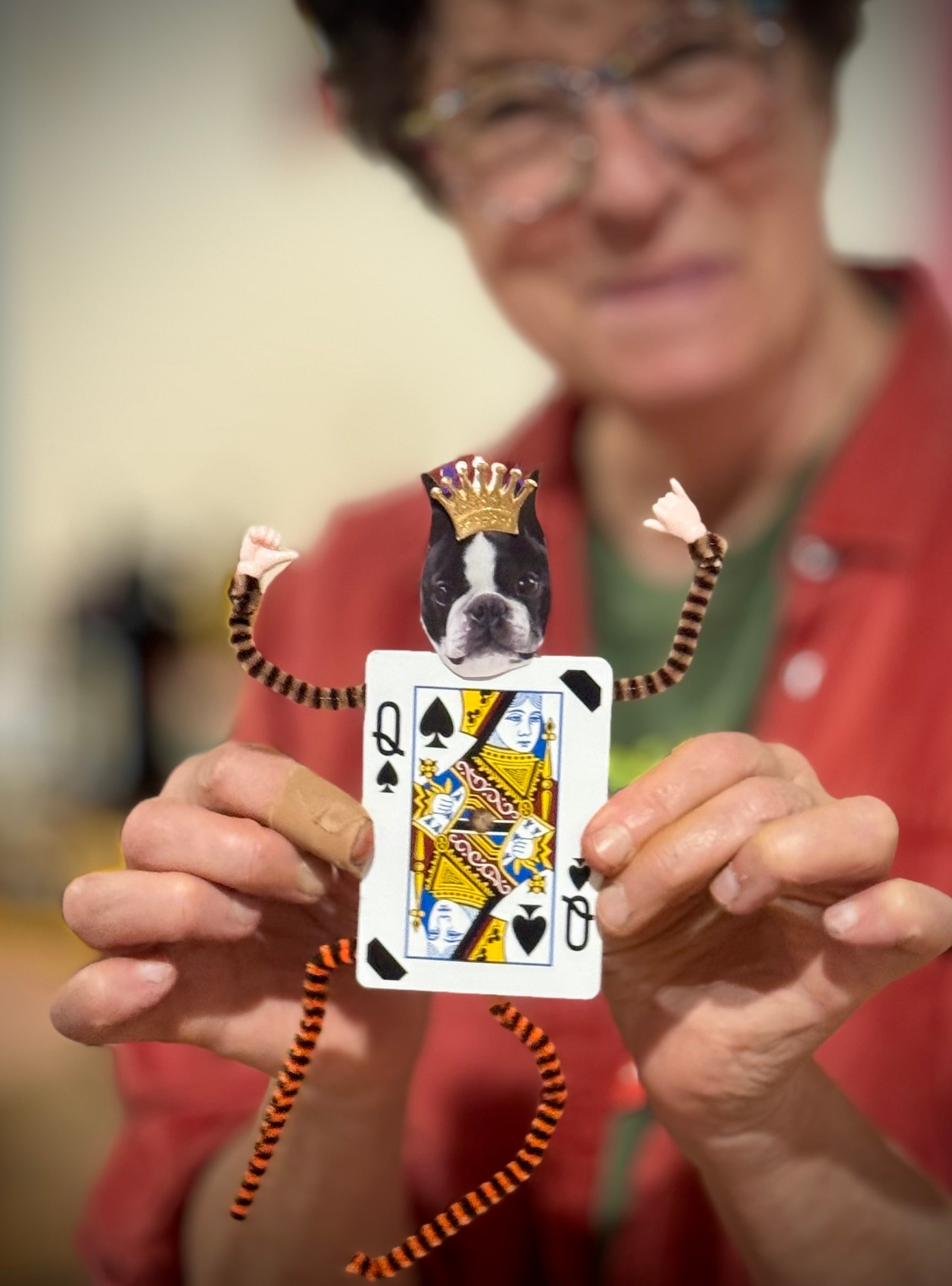 A Queen of Spades playing card turned into a little figure with a Boston terrier's head wearing a gold crown and striped pipe cleaner arms and legs, held up by two hands.