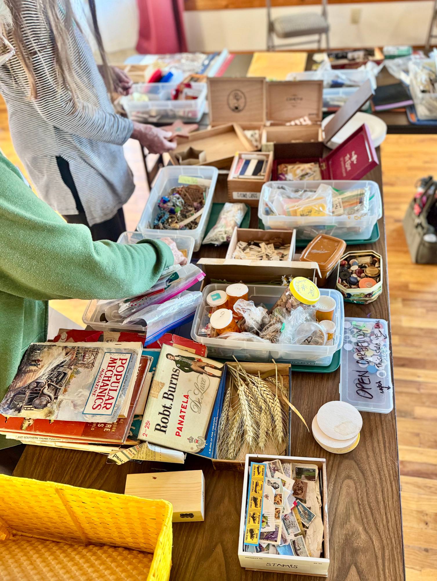 A long table seen from above, crowded with cigar boxes, tins of buttons, a box of postage stamps, old magazines and paperbacks, dried wheat, and plastic bins of craft supplies.